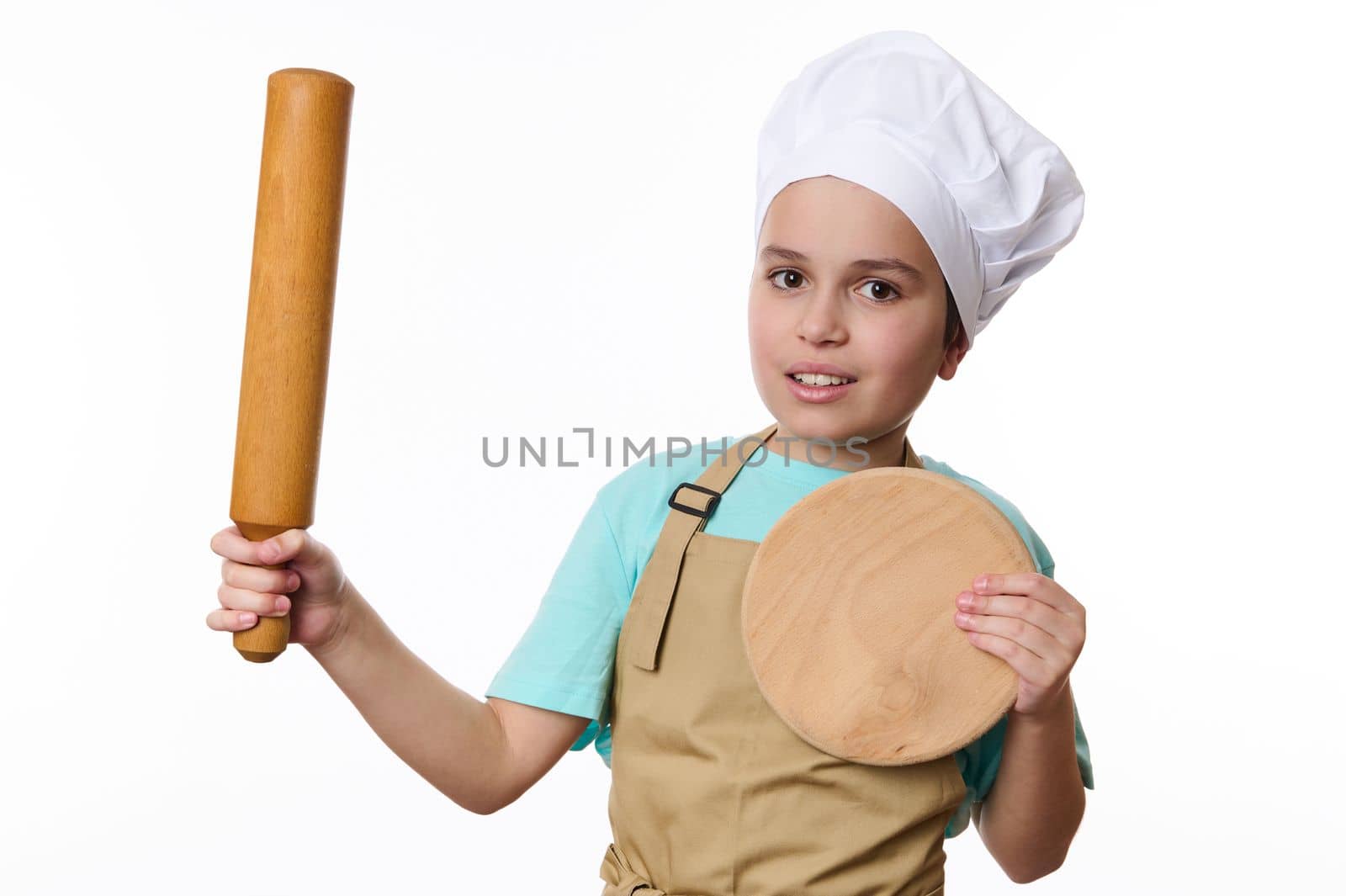 Adorable Caucasian child boy, wearing a chef's hat and beige apron, posing with a rolling pin and wooden cutting board, isolated on white background. Baker Confectioner. Chef pastry. Cook profession