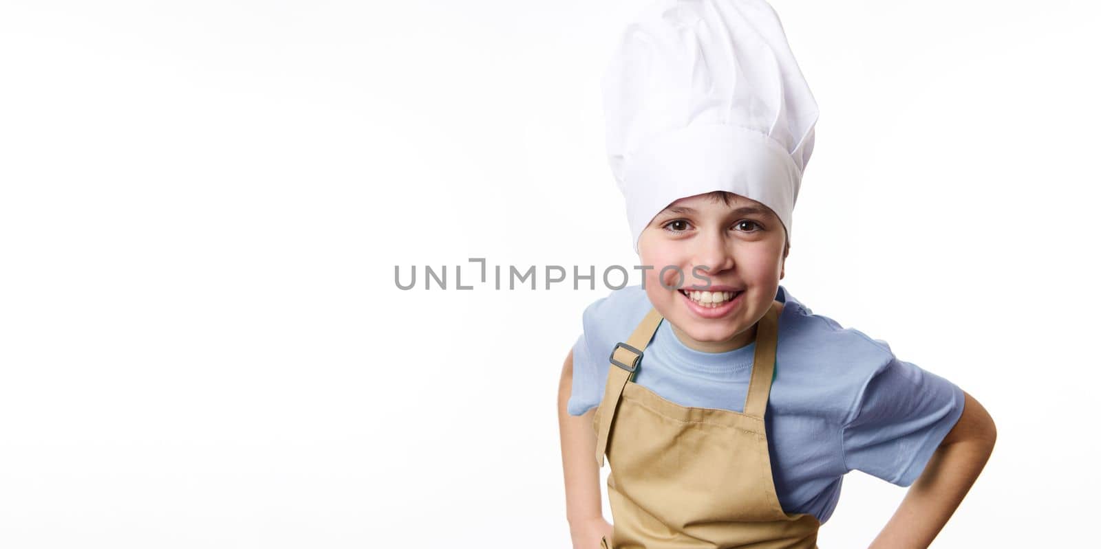 Horizontal studio shot on white background of delightful amazing schoolboy cook baker, wearing white chef hat and beige apron holding hands on belt, cheerfully smiling a toothy smile looking at camera