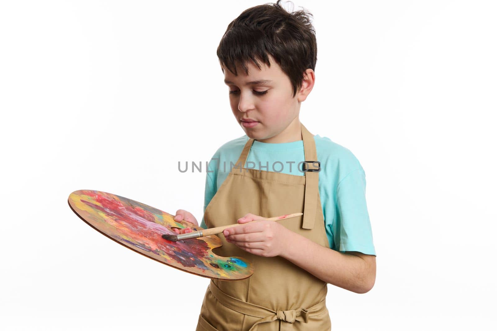Concentrated multi-ethnic teenage boy, inspired talented painter wearing beige apron, dipping paintbrush into a wooden palette with colorful paints, isolated on white background. Creativity ART class