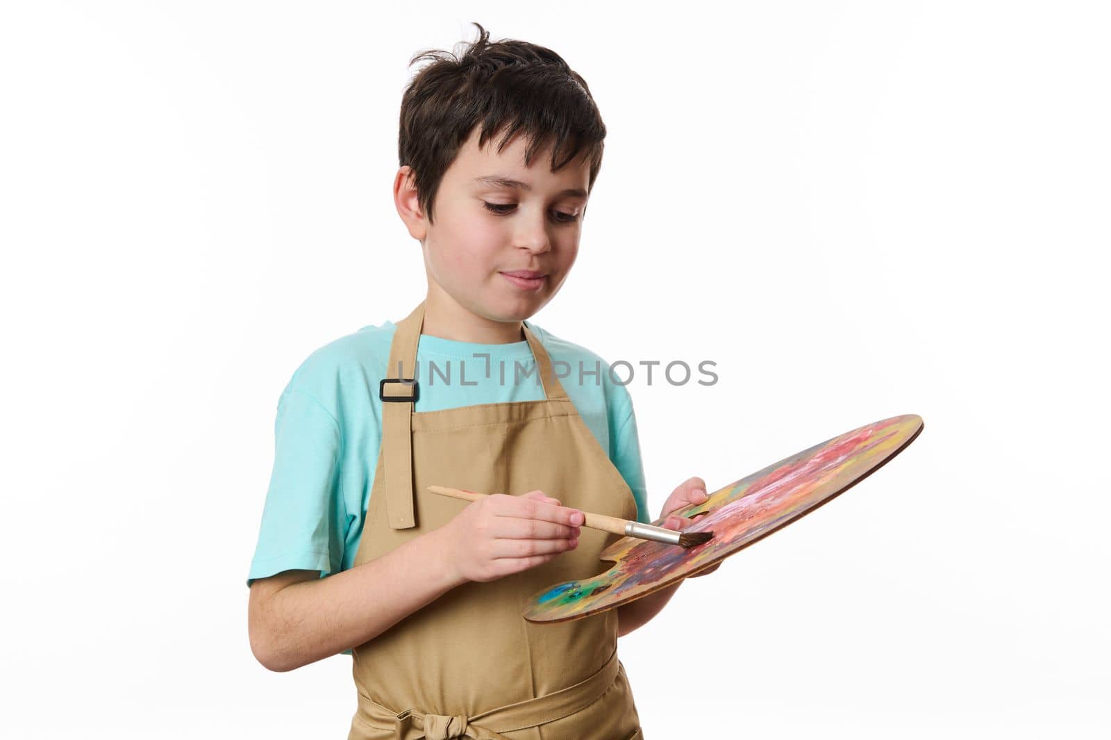 Inspired creative teenage boy, talented painter, artist wearing beige apron, dips a paintbrush on wooden palette with colorful acrylic, oil, watercolors paints, isolated on white background. Ad space
