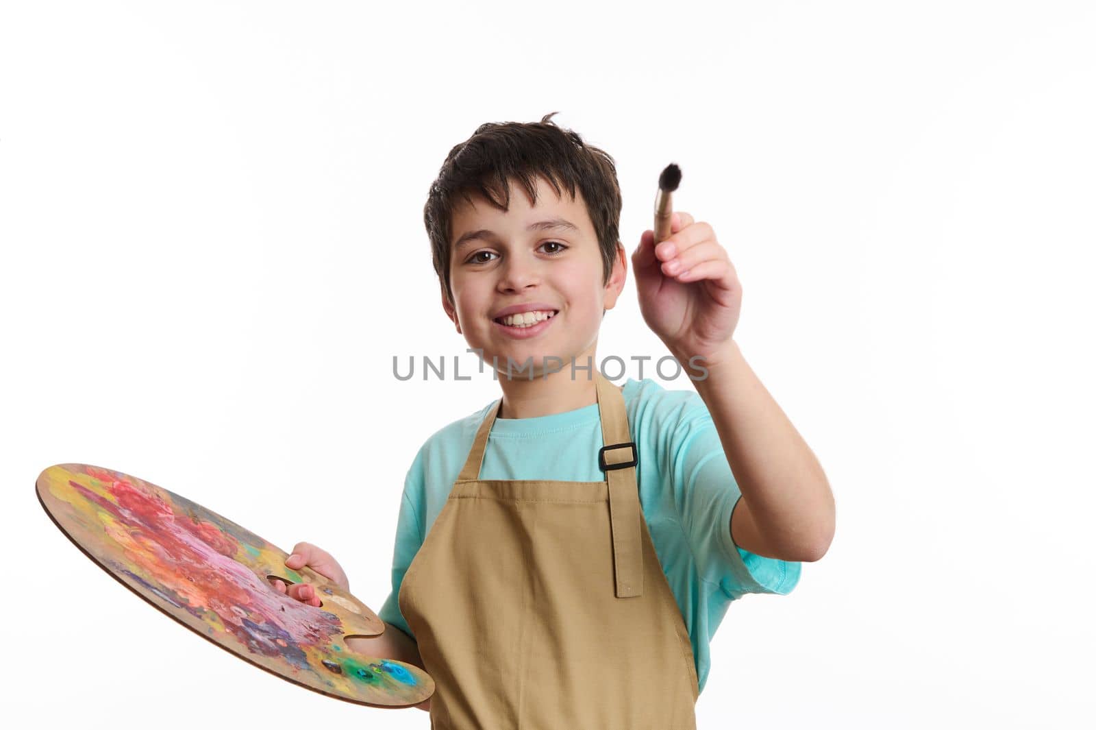 Artistic teenage boy wearing beige apron, holding a paintbrush and a color palette, paints on imaginary canvas, looking at camera, isolated on white background. Kids entertainment and art development