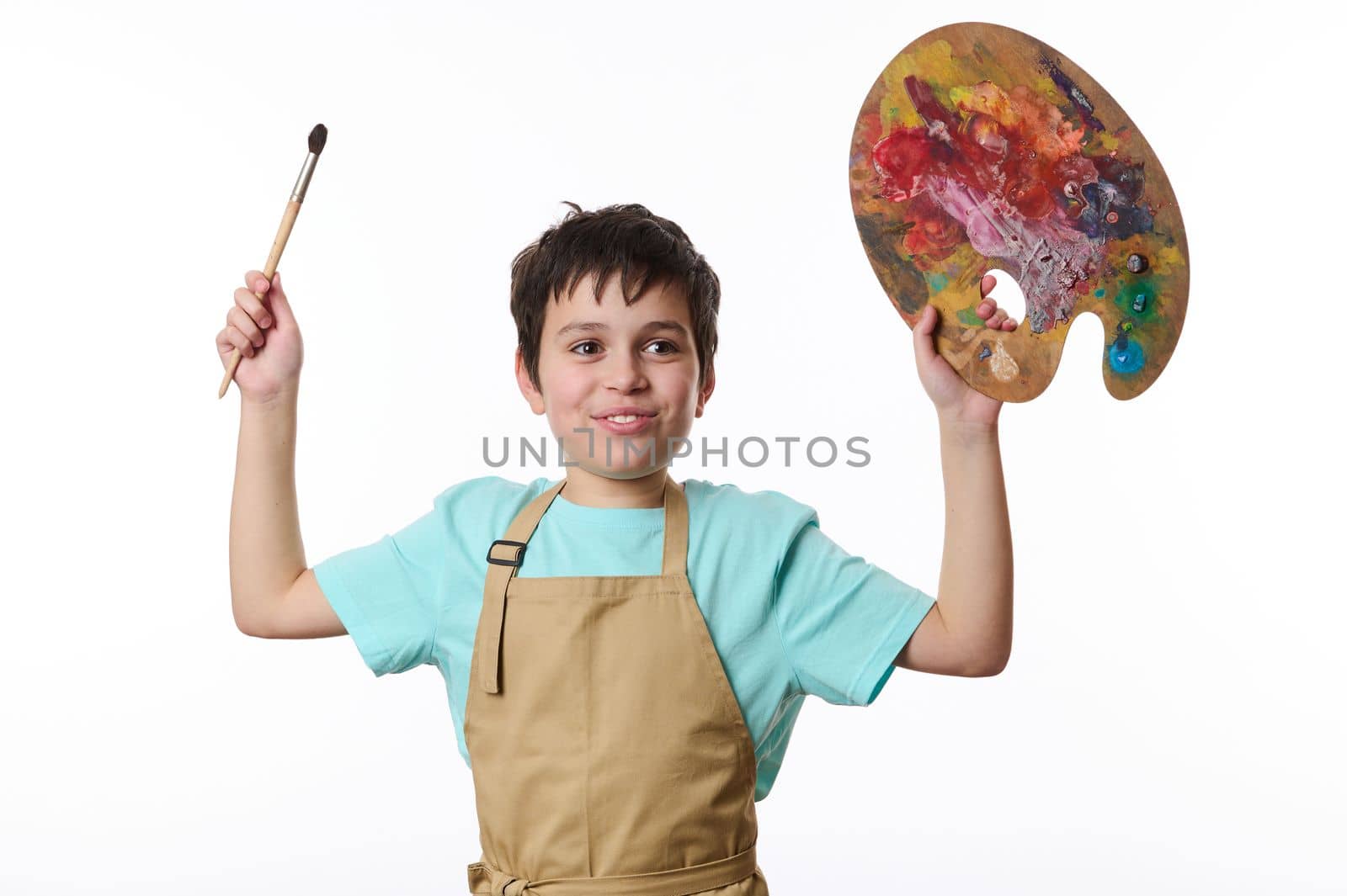 Inspired smiling teenage boy - talented painter in blue t-shirt and beige apron holding paintbrush and wooden palette, Development of children's creativity and drawing with paints. Art. Entertainment