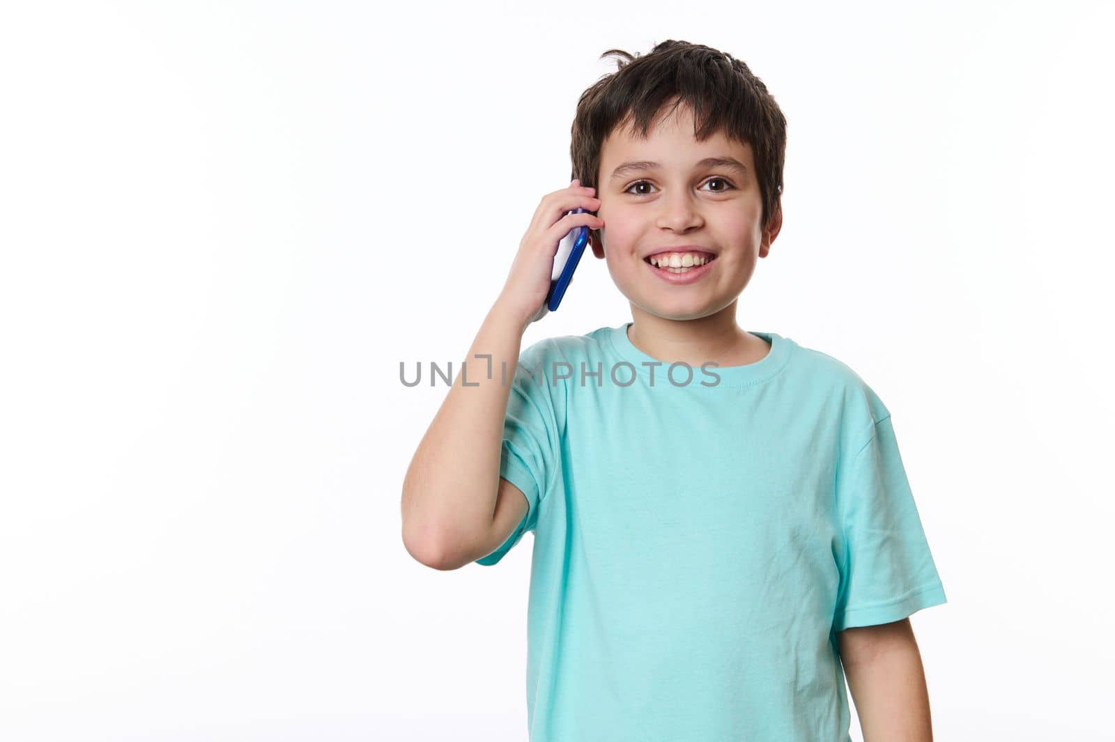 Overjoyed excited dark-haired Caucasian boy, wearing turquoise t-shirt, cutely smiling a cheerful toothy smile looking at camera while talking on mobile phone, over white background with copy space