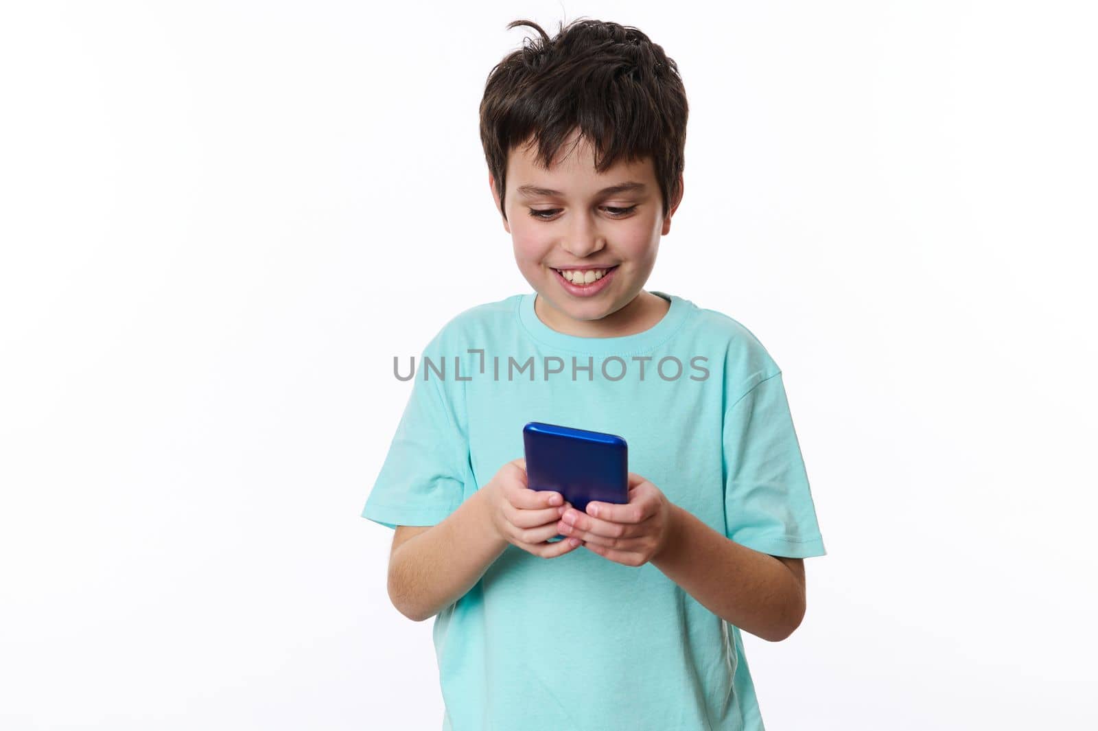 Isolated portrait on white background of a joyful happy teenage boy, wearing turquoise t-shirt, playing virtual online games on his smartphone. Digital gadget and entertainments for kids. Copy space
