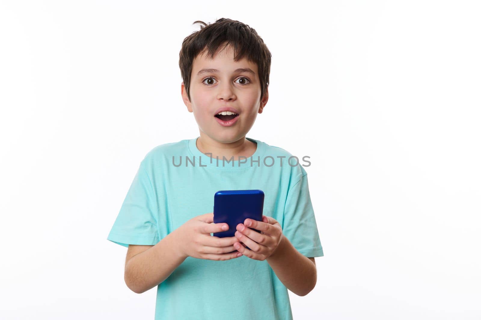 Amazed excited cute teenage boy, holding a cell phone and looking at camera with shocked face, child surprised by telephone, online service. Indoor studio shot white background. Wow awesome mobile app