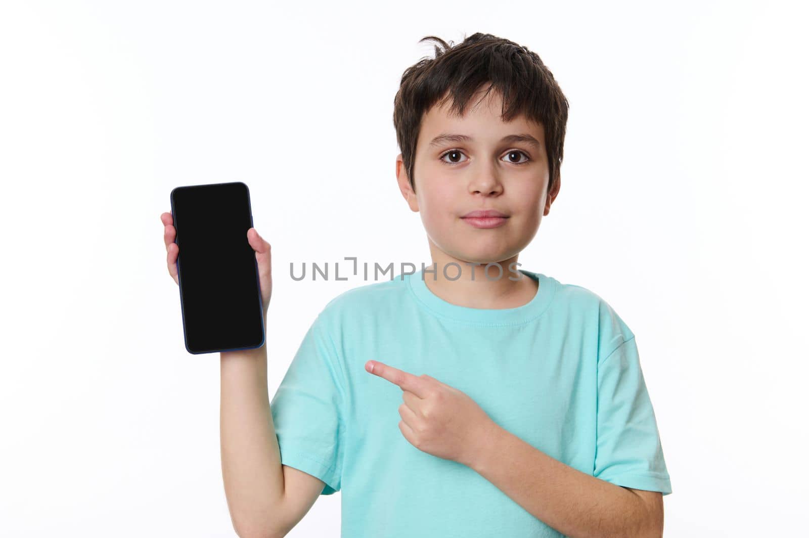 Isolated portrait on white background of adorable Hispanic preteen boy, wearing a blue t-shirt, pointing at a smartphone's blank screen with copy space for your promotional text or mobile application