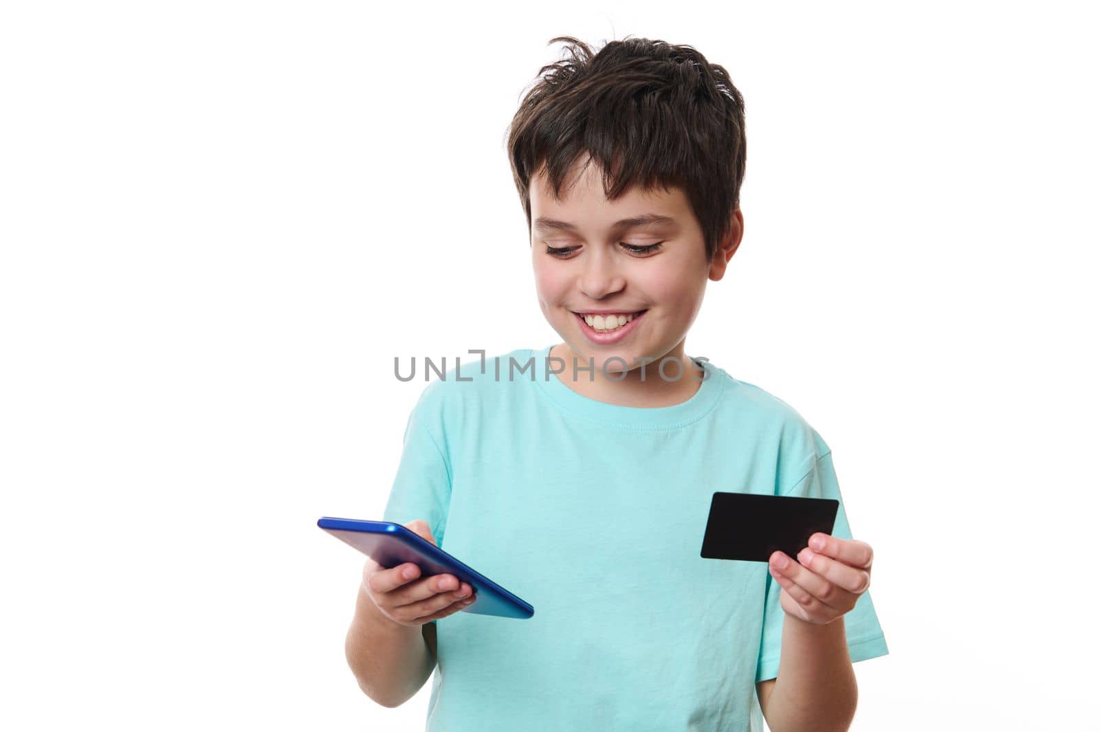 Isolated portrait on white background of a cheerful handsome smiling Caucasian teenage schoolboy, using mobile phone and credit card, makes cashless payment while shopping online, Internet banking