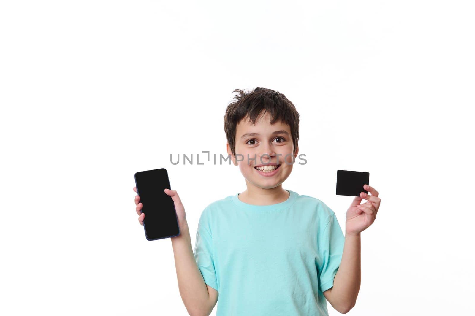 Adorable teenager boy smiles happily at camera, showing a smartphone and a black credit card with free space for advertising text, isolated on a white background. Internet banking. Mobile apps concept