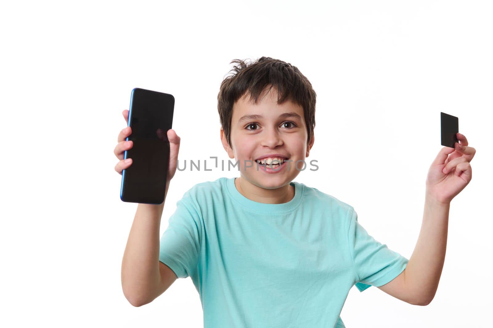 Happy overjoyed handsome preteen boy holding smartphone and ATM card, with advertising space for promotional text and mobile application, isolated on white background. Internet banking Online shopping