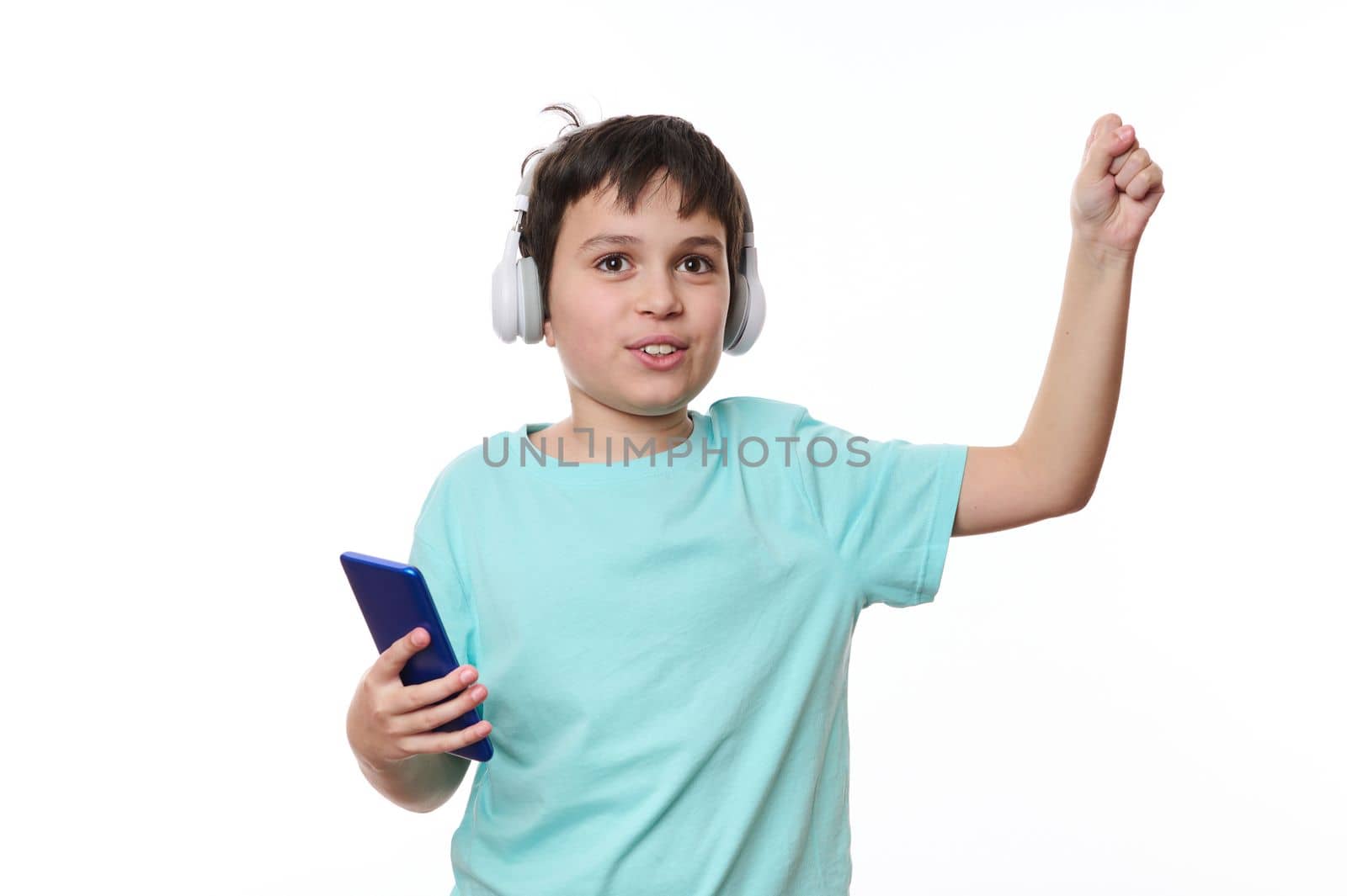 Adorable child, teenage boy listening to the music on wireless stereo headphones, adjusting tracks on his smartphone, isolated over white background with copy advertising space for promotional text