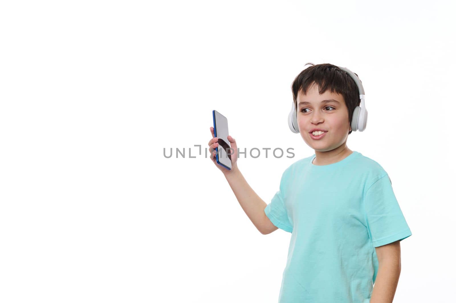 Lovely child, teenage boy wearing blue turquoise t-shirt, holding smartphone and dancing while listening to the music on stereo headphones, isolated over white background with copy ad space for text