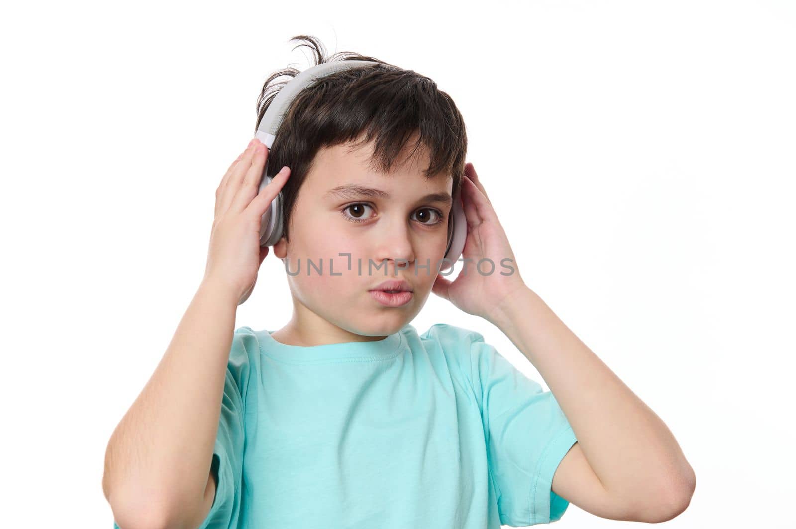 Close-up portrait handsome multi-ethnic teenage boy in blue t-shirt, putting on wireless stereo headphones, looking at camera, isolated on white background, copy advertising space for promotional text