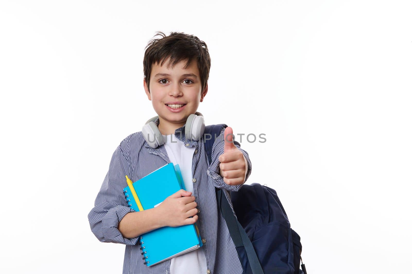 Caucasian smart primary school student, teenage boy in casual wear, with wireless headphones, backpack and school supplies, thumbs up looking at camera, white background. Copy ad space. Back to school