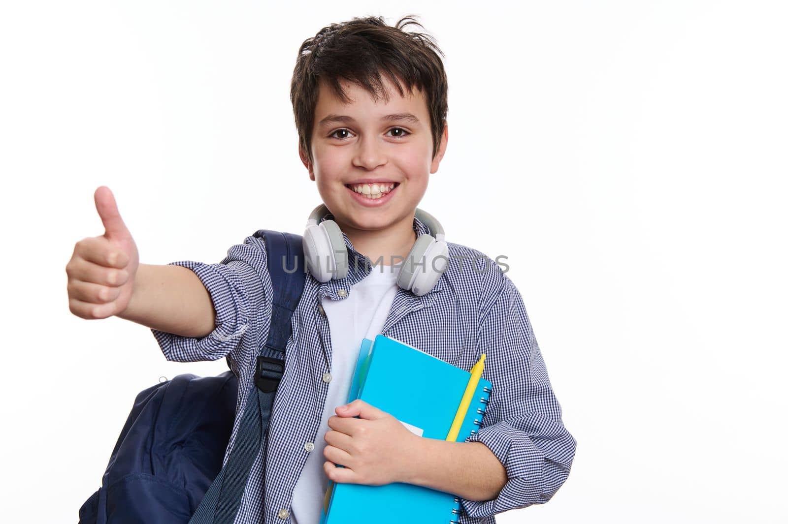 Caucasian teenage schoolboy with backpack and copybook, smiling a cheerful toothy smile and showing thumb up at camera, isolated on white background. Back to school. Knowledge. Education concept