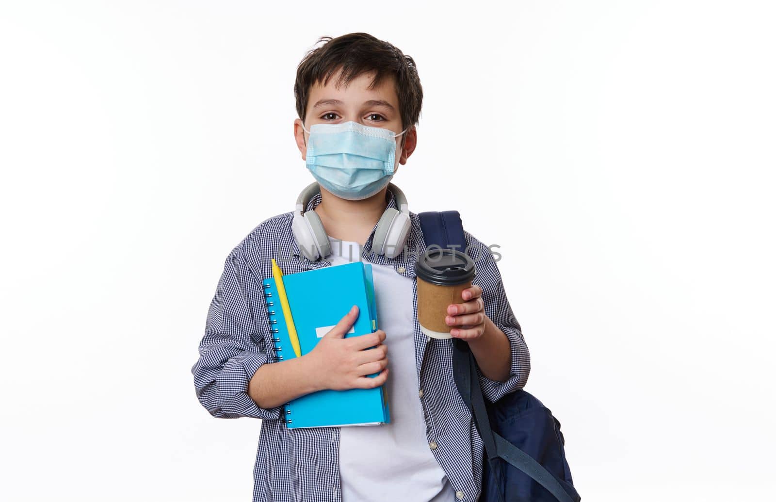 Adorable positive teen boy, primary school student with schoolbag and copybook, holding out a disposable eco paper cup with hot drink at camera, isolated on background. Copy space