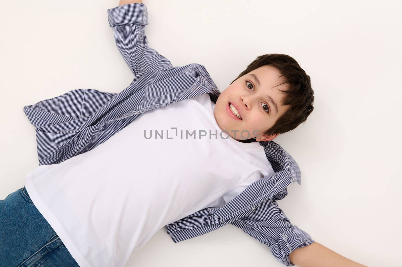 View from above of Caucasian adorable teen boy in blue jeans, checkered shirt and white t-shirt, looking at camera, lying down on a white background. Copy advertising space. Portrait of beautiful kids