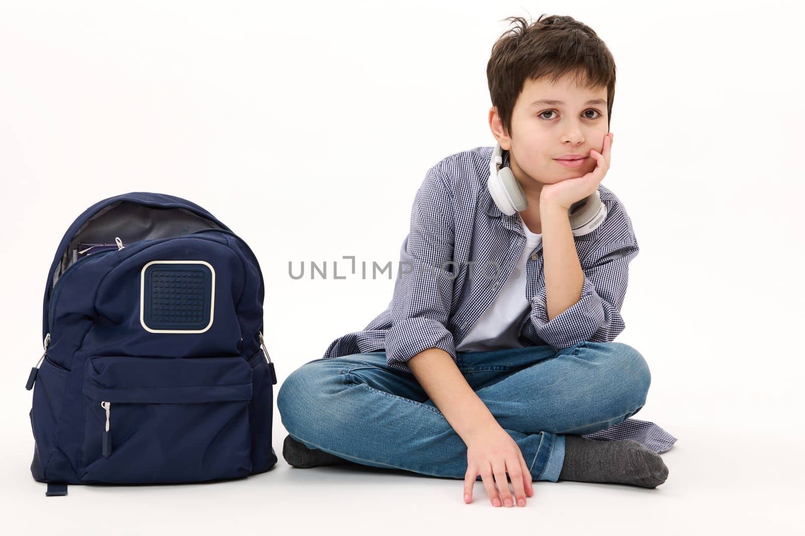 Lovely Caucasian boy teenager, elementary school student wearing casual denim, with blue backpack, confidently looking at camera, isolated on white background. Copy advertising space. Back to school