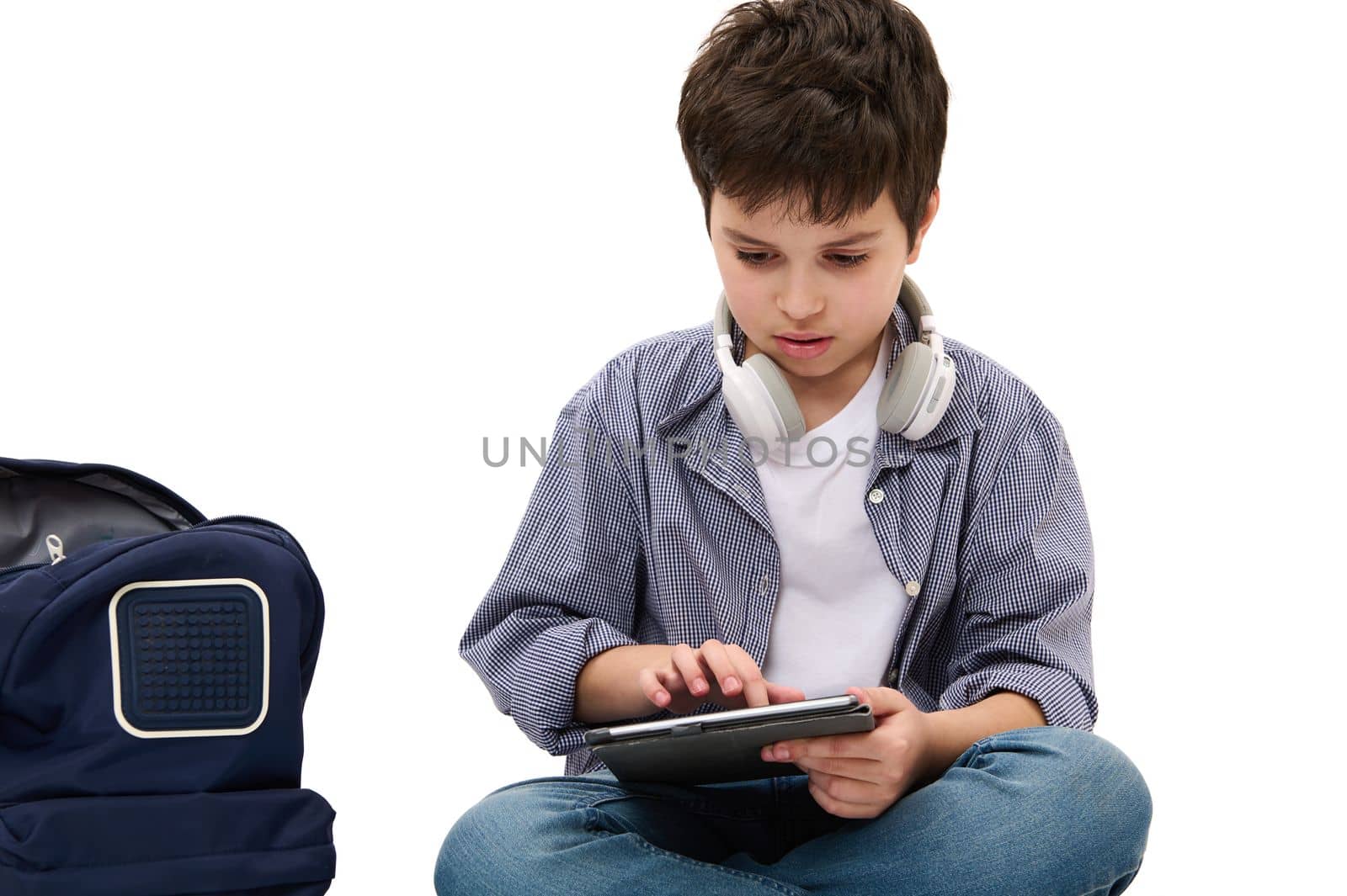 Caucasian smart elementary age student, teen boy in casual denim working on his school project using digital tablet, sitting near his backpack, isolated over white background. Copy advertising space