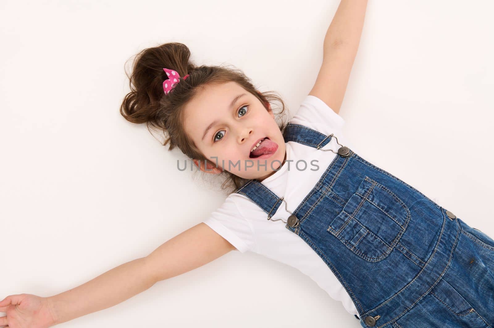 View from above of a Caucasian mischievous child, a lovely baby girl in blue denim overalls and white t-shirt, lying down on a white background. Copy advertising space. Beautiful kids.