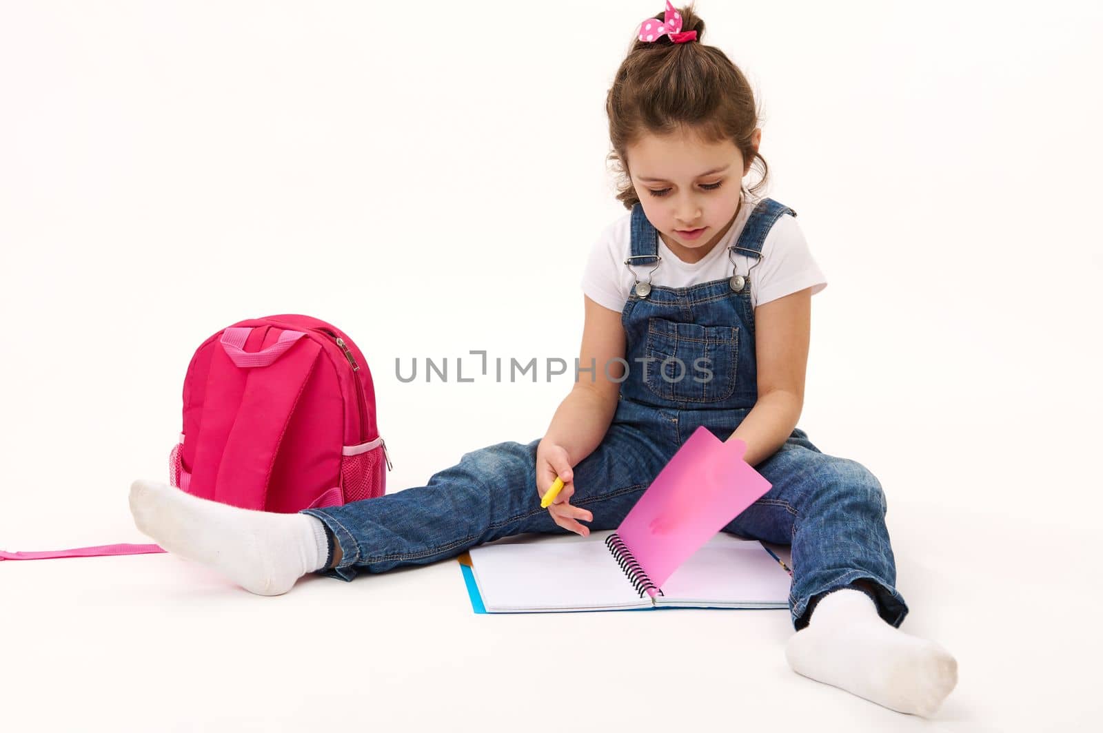 Charming fist grade student, adorable baby girl in white t-shirt and blue denim overalls, sitting near her pink backpack and copybook, isolated over white background. Back to school on new semester
