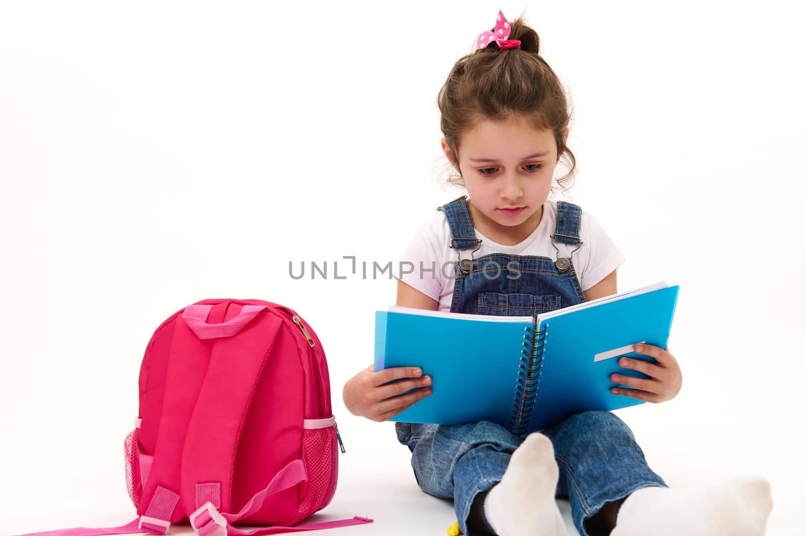 Caucasian lovely child, little girl in blue denim overalls and t-shirt, first grade student reading textbook, sitting near her pink backpack on white background. Copy ad space. Back to school concept