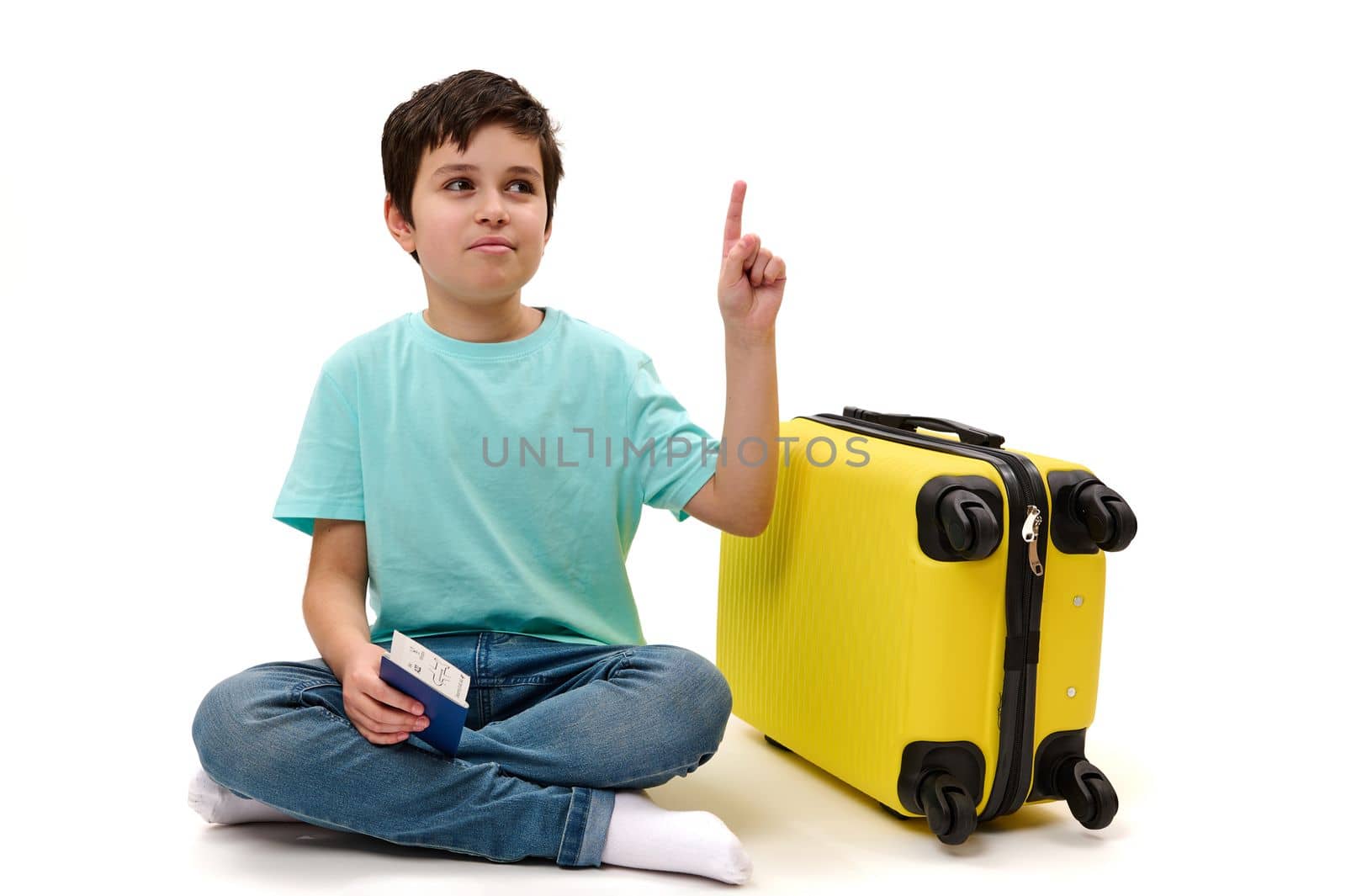Adorable preteen child, teenage traveler boy in blue denim jeans and casual t-shirt, holding a boarding pass, pointing with index finger at ad space, sitting near yellow suitcase over white background