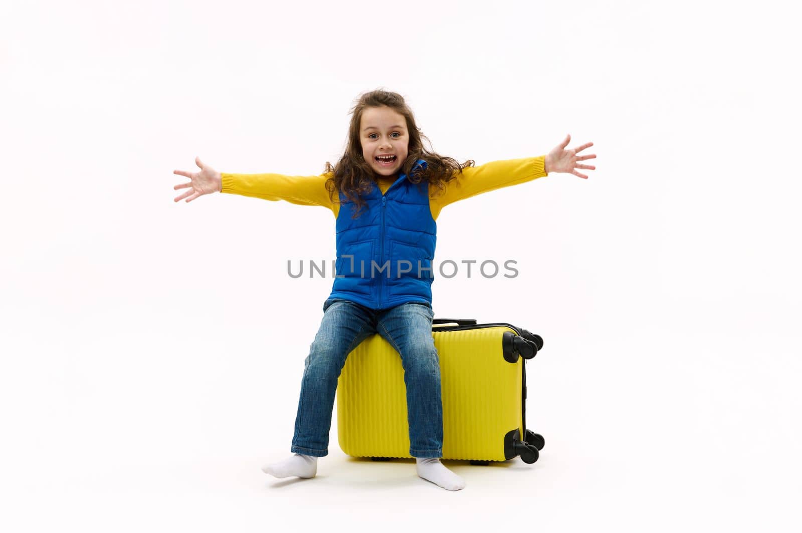 Lovely little girl in blue jacket looks at camera, sitting on yellow suitcase with outstretched arms on white background by artgf