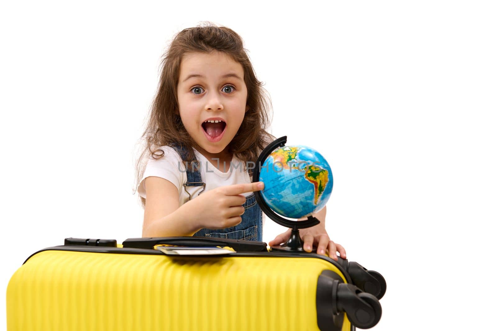 Cute little girl traveler with a yellow suitcase points to a destination on the globe, looking at the camera in surprise by artgf