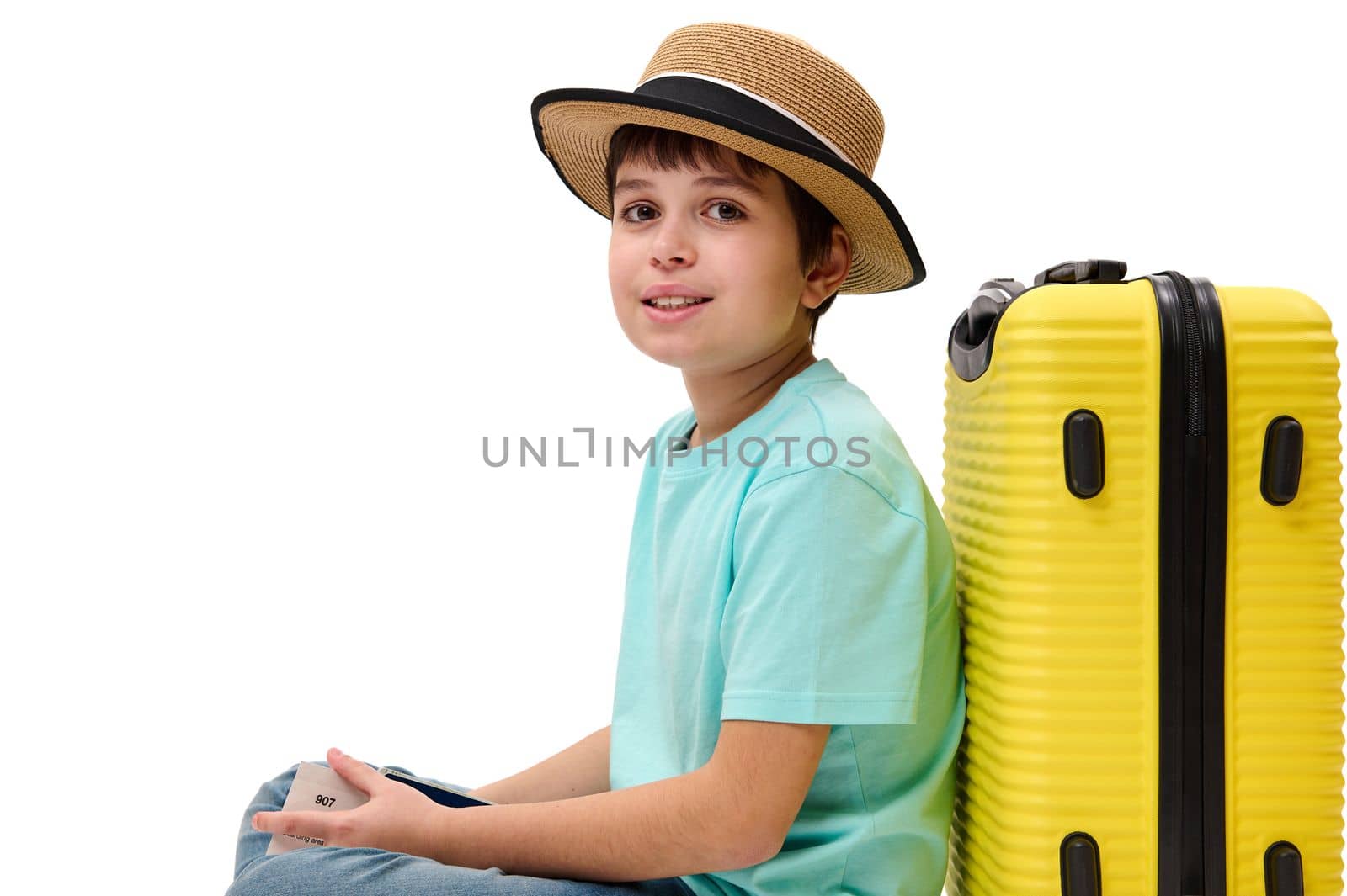 Multi-ethnic charming teenage boy in turquoise t-shirt and straw hat, posing with suitcase and boarding pass, looking at camera, isolated over white background. Traveling, holidays, children concept