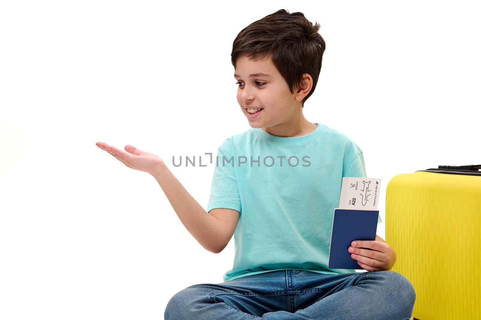 Teenage traveler boy in casual wear with boarding pass, sitting next to yellow suitcase, looking at imaginary advertising space on his hand palm up isolated on white background. Tourism Travel concept