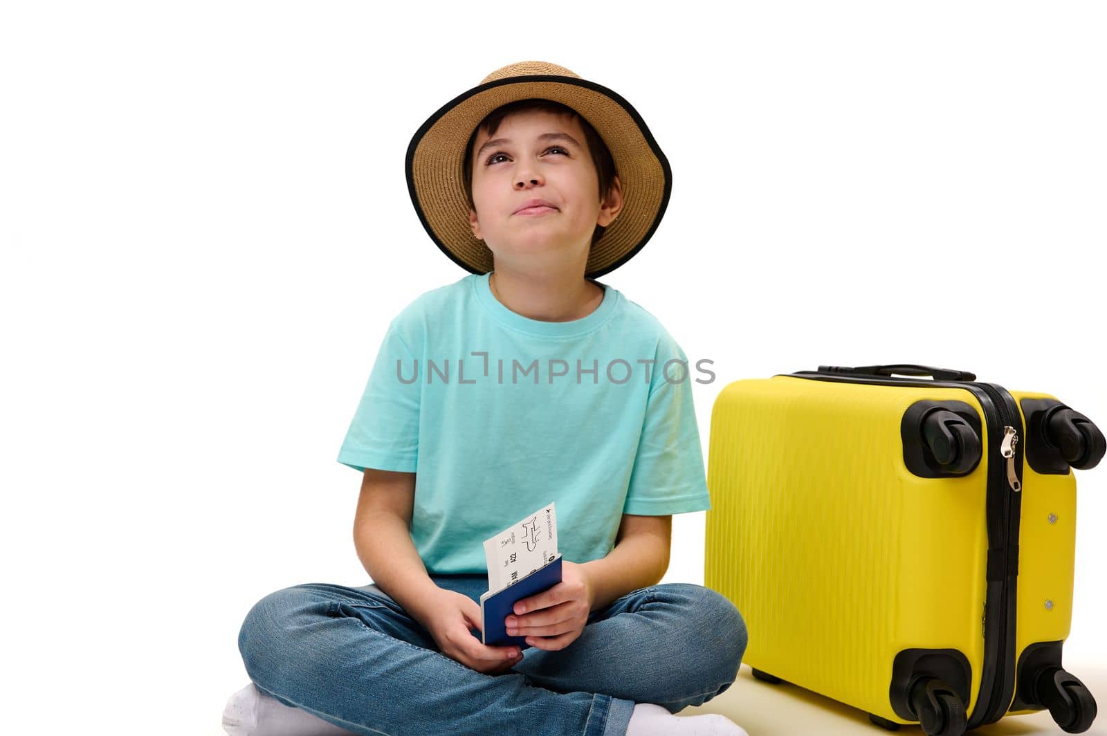 Isolated portrait on white background of teenage boy traveler in casual wear, with boarding pass and suitcase, dreamily looking aside, traveling abroad for the weekend. Tourism and air travel concept