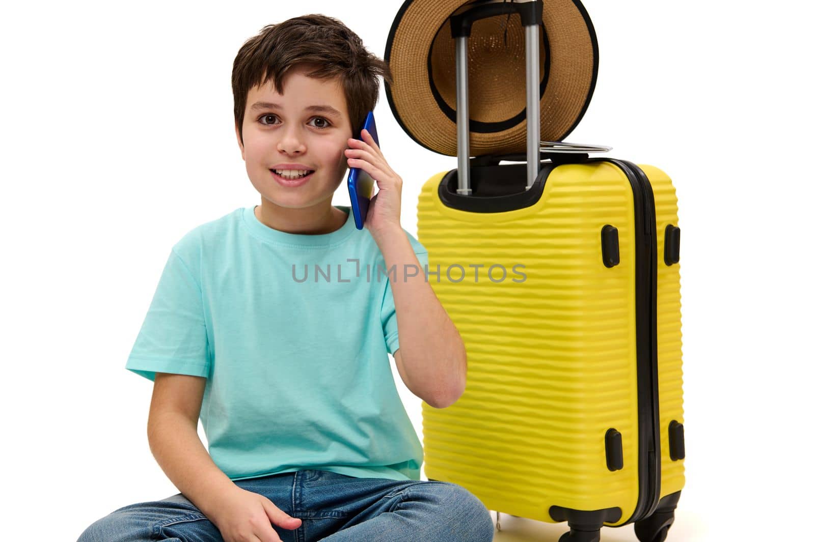 Lovely dark-haired Caucasian teen boy, wearing a turquoise t-hirt and blue casual jeans, looking at camera while talking on mobile phone, sitting over yellow suitcase on white background. Copy space