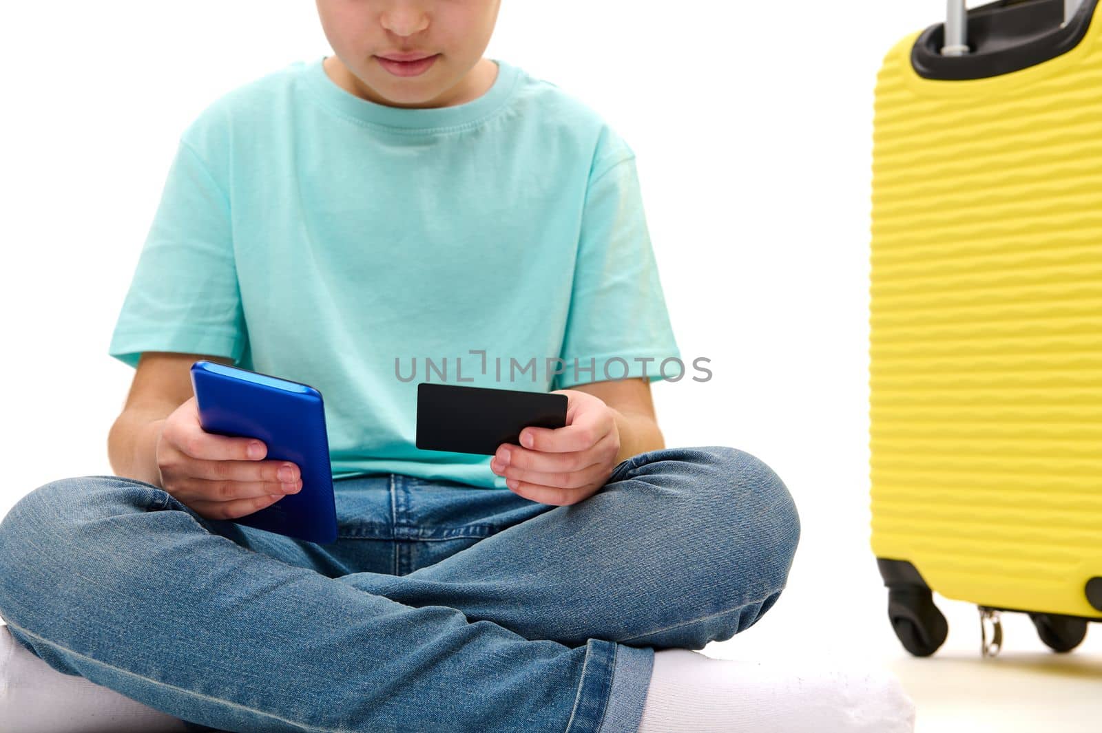 Cropped view of a traveler teenage boy making online payment, using credit card and modern smartphone, sitting near yellow suitcase on white background. Internet banking. Cashless payment concept