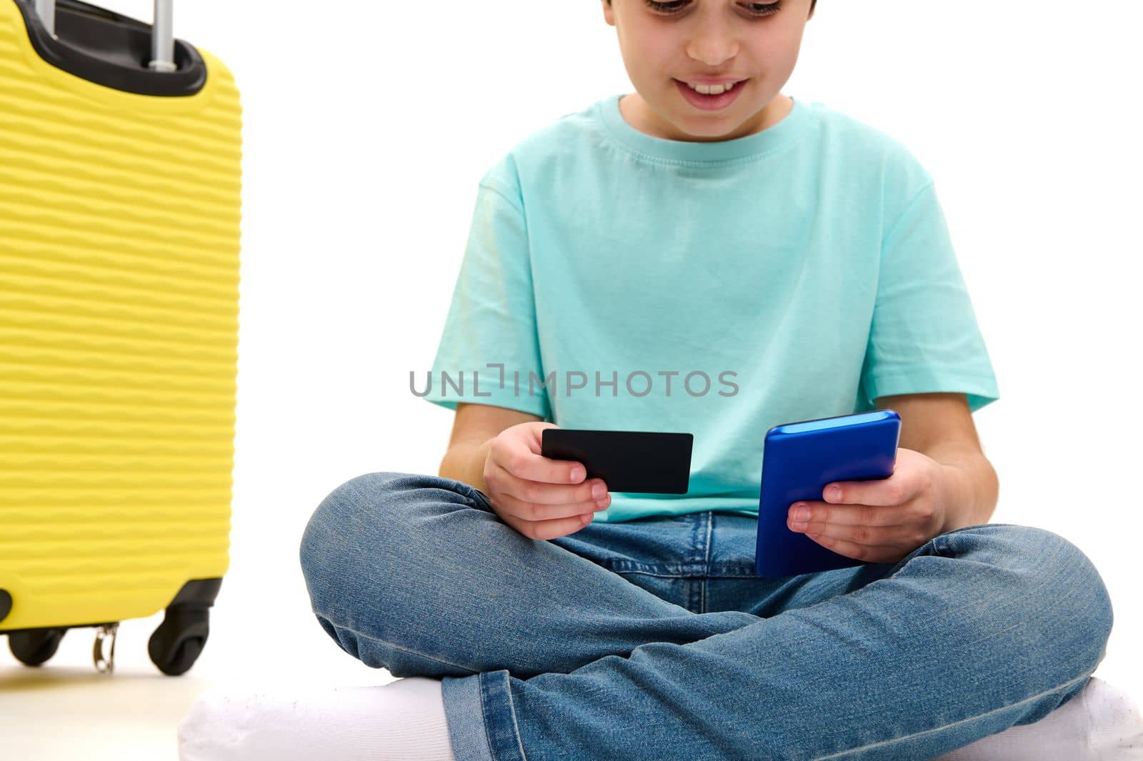 Selective focus on the hands of a teenage boy holding modern smartphone and credit card, making online payment, sitting near yellow suitcase over white background with copy space for your advertising