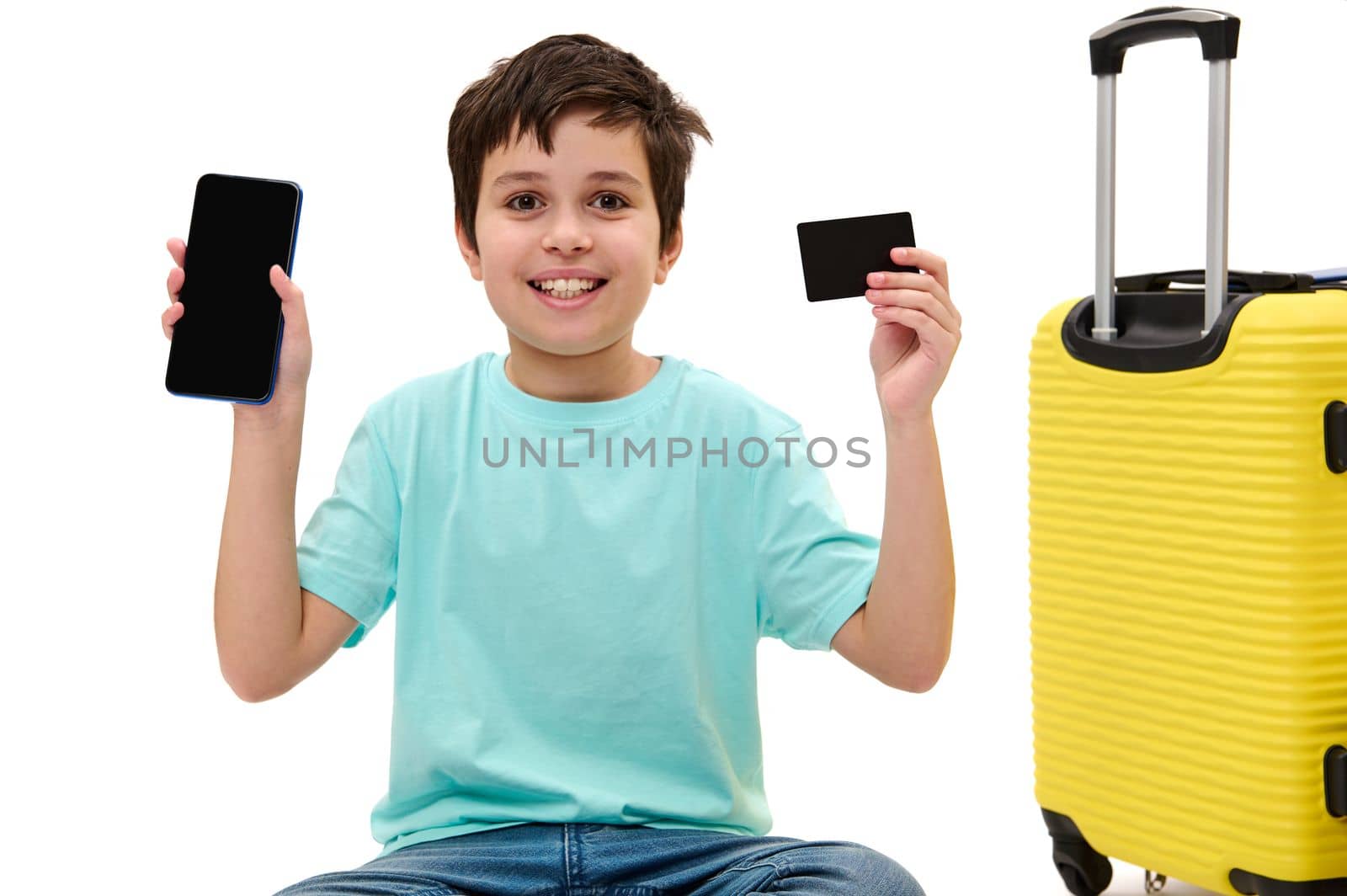 Adorable Caucasian child boy in blue jeans and t-shirt, looking at camera, showing his smartphone and plastic bank card with copy advertising space to camera, sitting near suitcase over white backdrop
