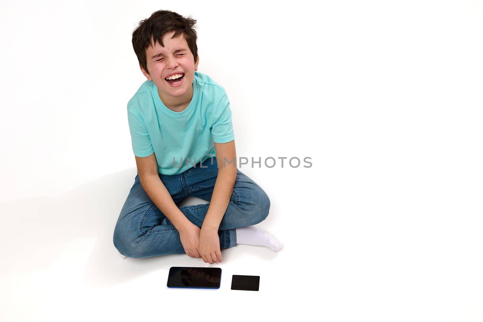View from above of dissatisfied Caucasian boy, in blue t-shirt and jeans, looking up, expressing sadness and frustration, sitting on white background with modern smartphone and credit card. Copy space