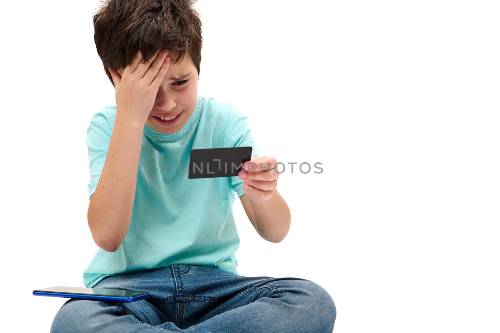 Dissatisfied Caucasian teenage boy in blue t-shirt, looking at his credit card, expressing disappointment and frustration while failing transaction, isolated on white background. Unsuccessful payment