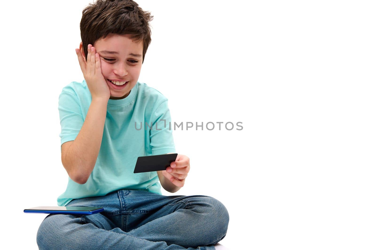 Upset Caucasian teenager in blue t-shirt, looking at his credit card, expressing shock and sadness at a failed transaction due to lack of money in his banking account, white background. Failed payment