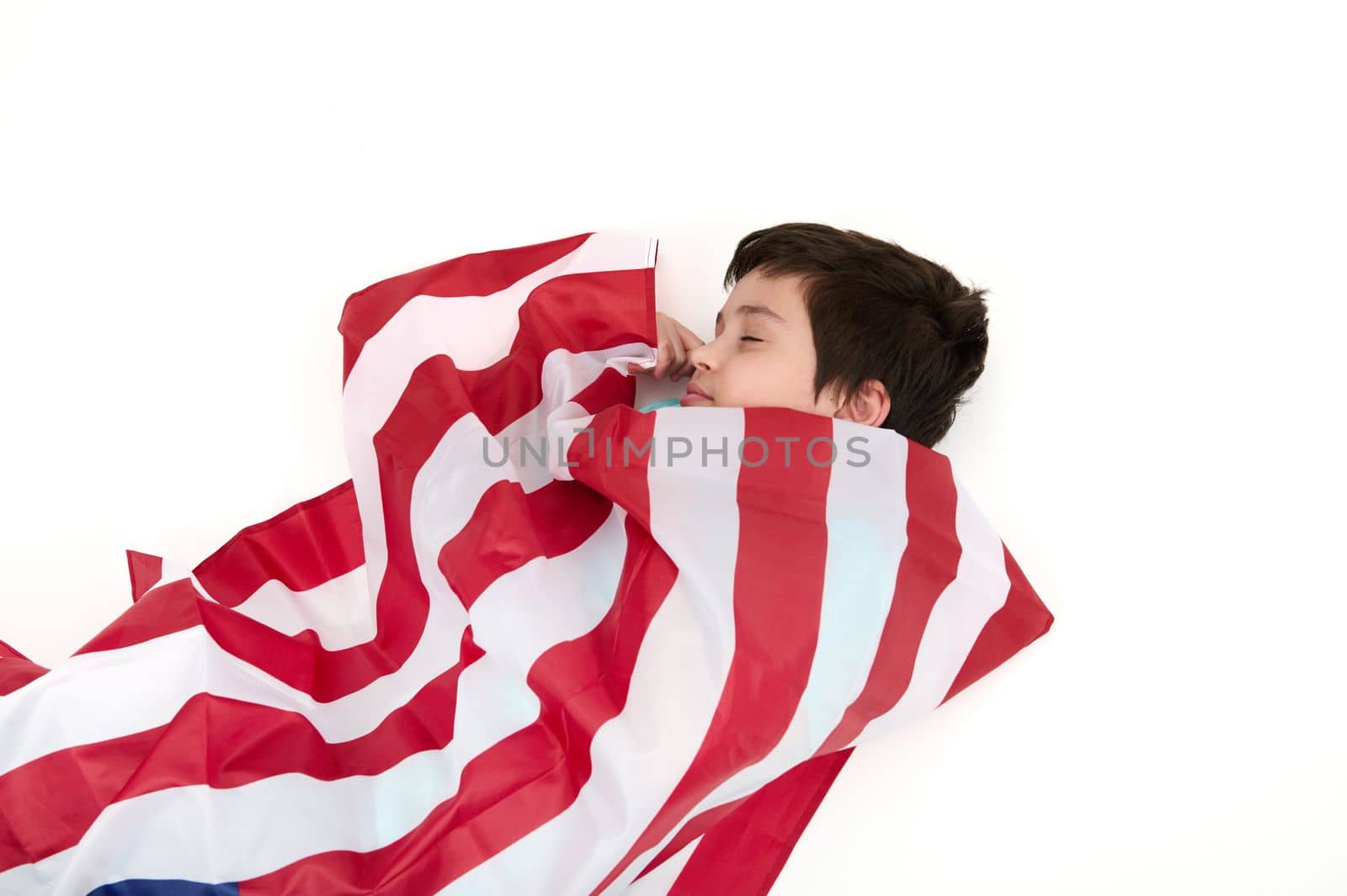 Serene multi-ethnic child boy wrapped in USA flag, isolated on white background. Concept of love for the motherland, citizenships, social secure, patriotism and freedom. Independence Day on July 4th