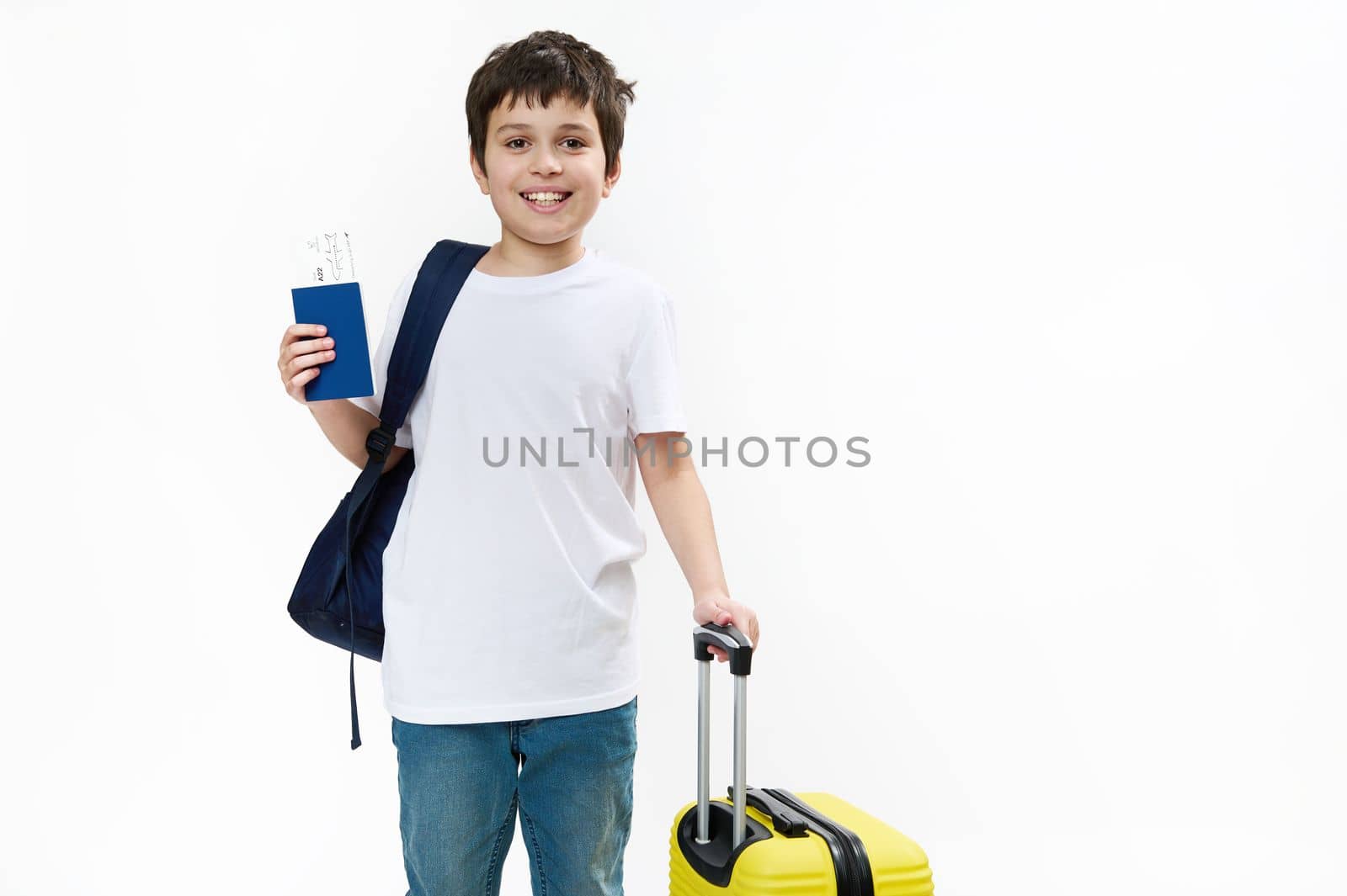 Happy teenage traveler tourist boy in blue jeans and casual t-shirt with backpack, yellow travel bag and boarding pass, smiling a cheerful toothy smile looking at camera, isolated on white background