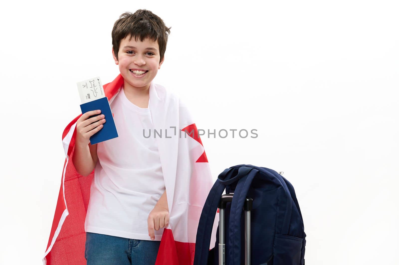 Multiethnic happy smart teenage boy in casual t-shirt and blue jeans, smiling, looking at camera, posing with Canadian flag, boarding pass and backpack on white background. Free space for advertising