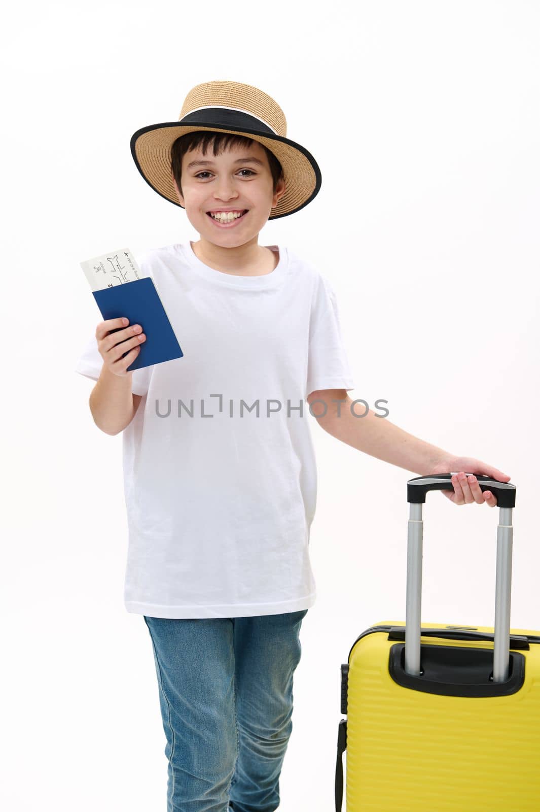 Happy preteen child passenger, teenager traveler boy in casual t-shirt, blue jeans and straw hat, going on summer holidays, walks with boarding pass, air ticket and yellow suitcase on white background