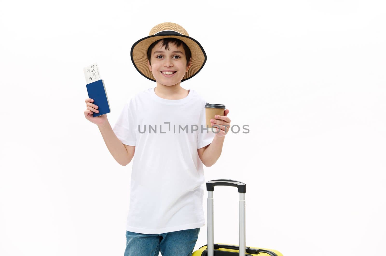 Happy teenage traveler tourist boy in blue jeans, casual t-shirt and straw hat with travel bag, holds a takeaway hot drink in disposable cardboard cup and boarding pass, isolated over white background