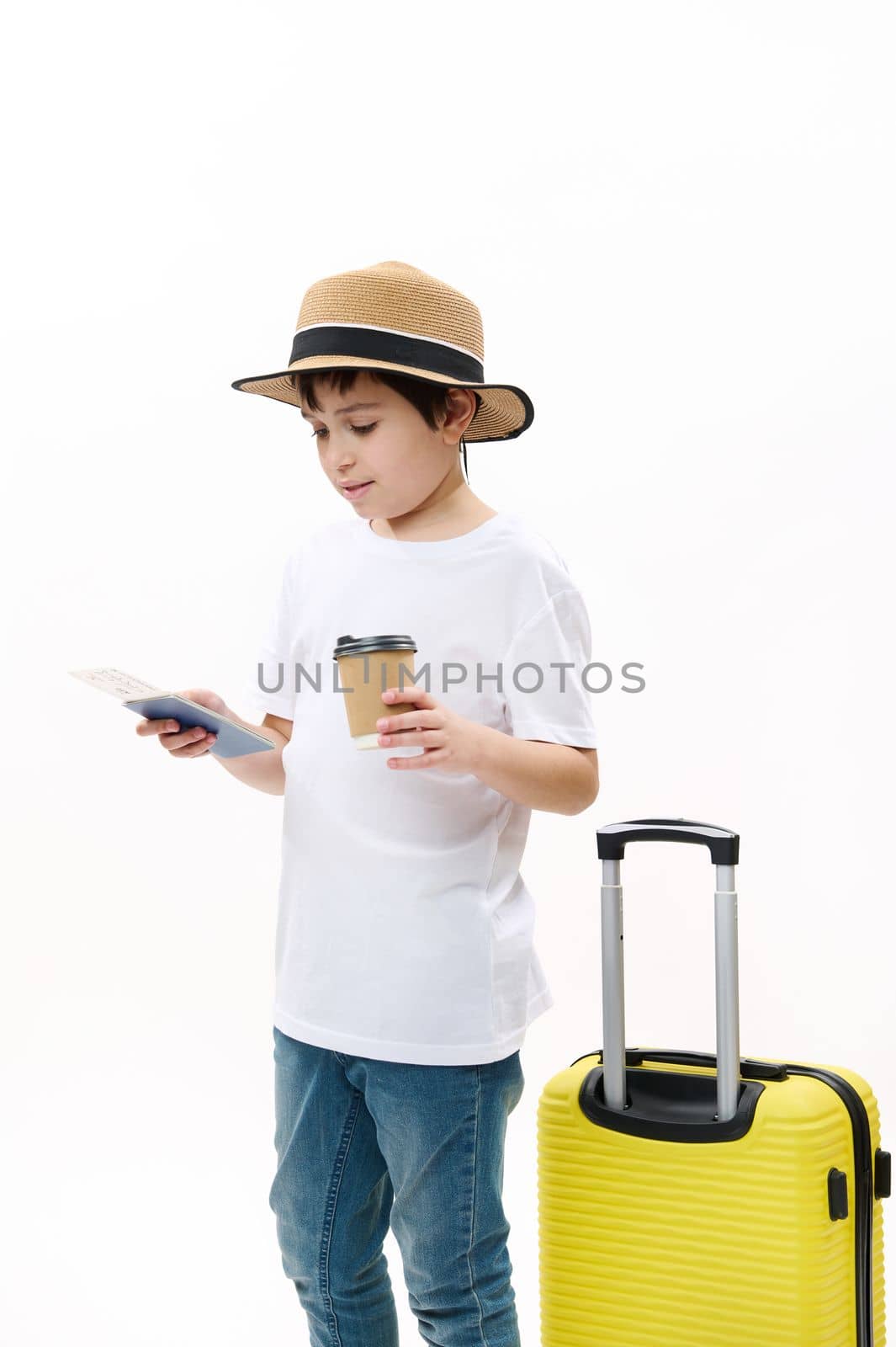 Adorable teenage boy in blue jeans, casual t-shirt and straw hat with yellow suitcase, holds a takeaway hot drink in disposable paper cup and looks at his air ticket, isolated over white background