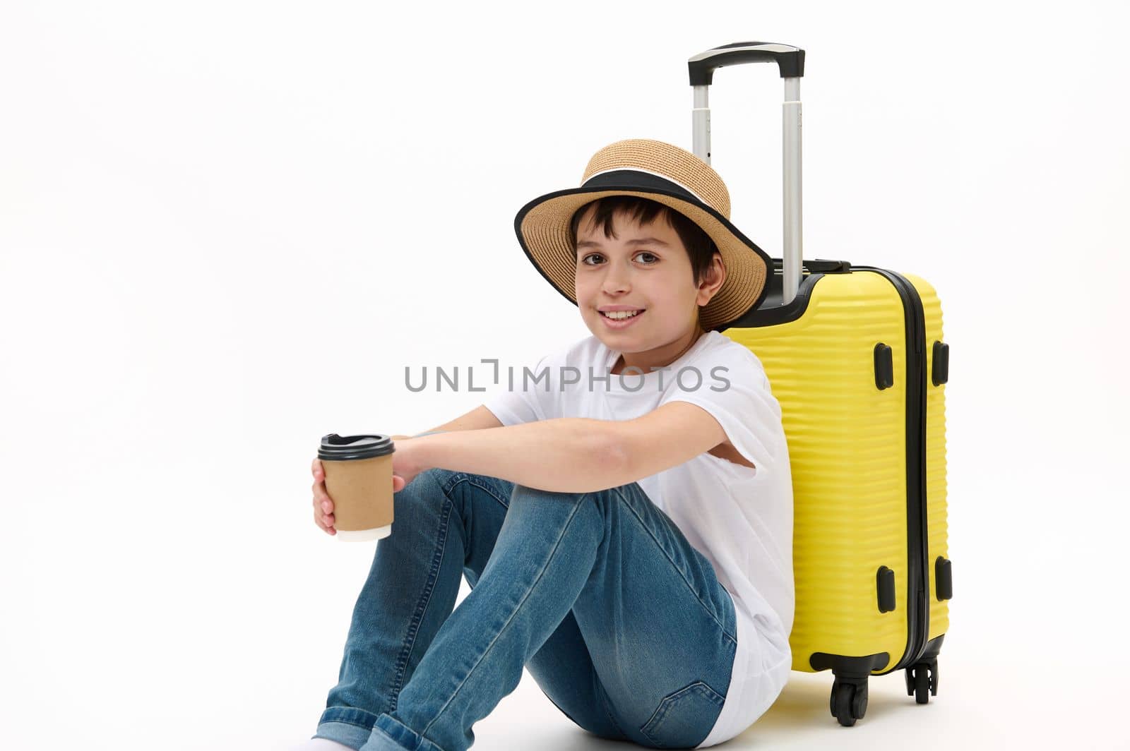Happy preteen boy traveler in casual wear and straw hat, with yellow suitcase and takeaway drink in disposable eco paper cup, waiting to board the flight, isolated on white background. Free ad space.