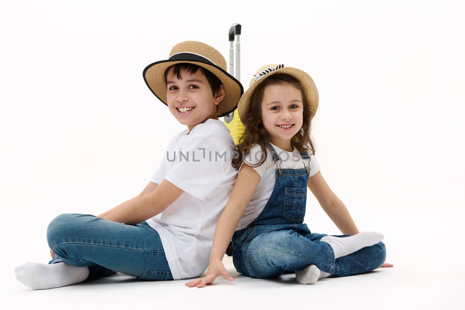 Adorable diverse children, preteen boy and little girl sit with their backs to each other against yellow suitcase, cutely smile on camera, isolated on white background. Kids traveling. Family journey