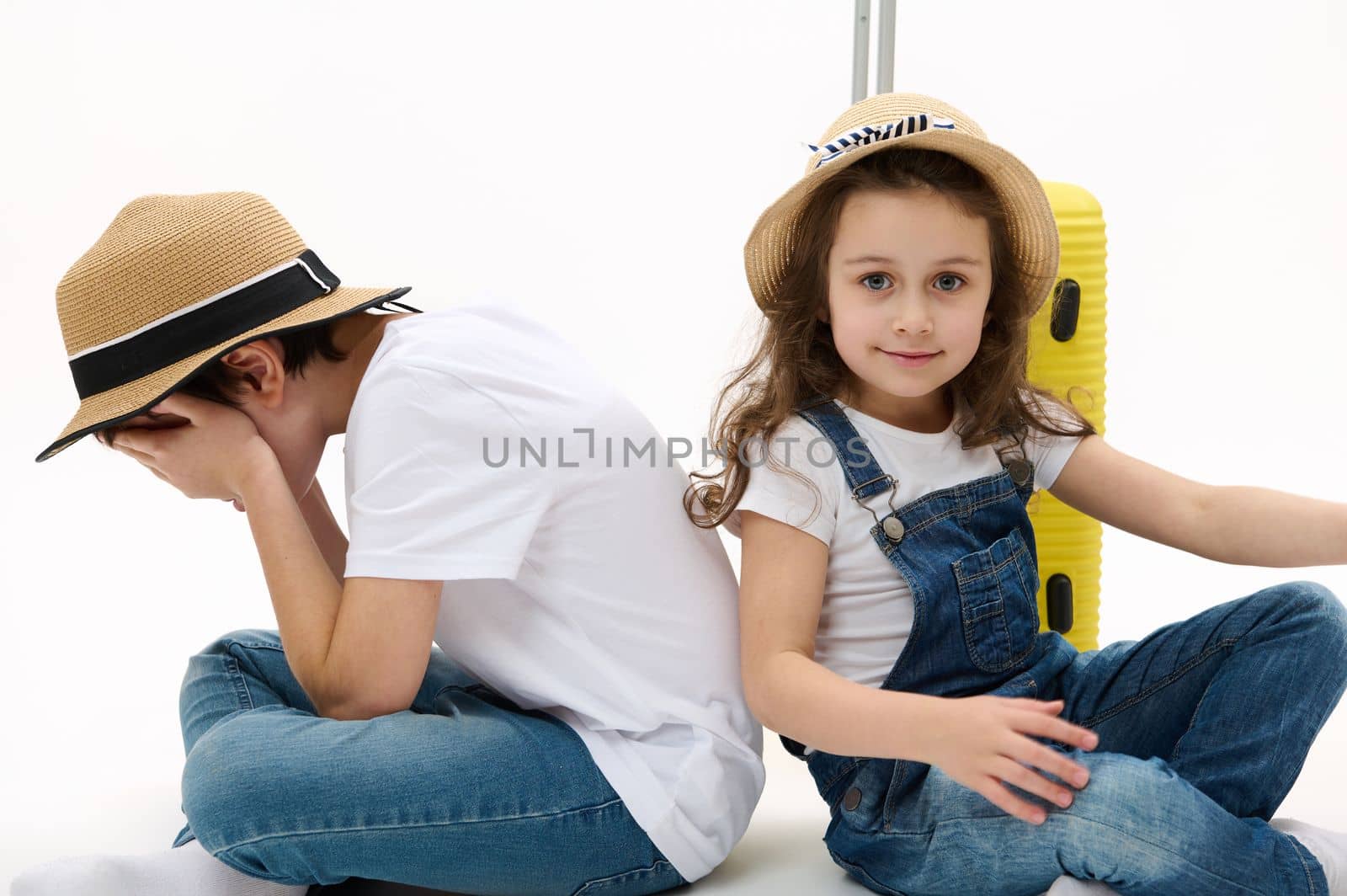 Beautiful Caucasian little girl wearing casual denim overalls and straw hat, looking at camera while sitting near her brother who hides his face, waiting to board the flight. Kids tourists travelling.