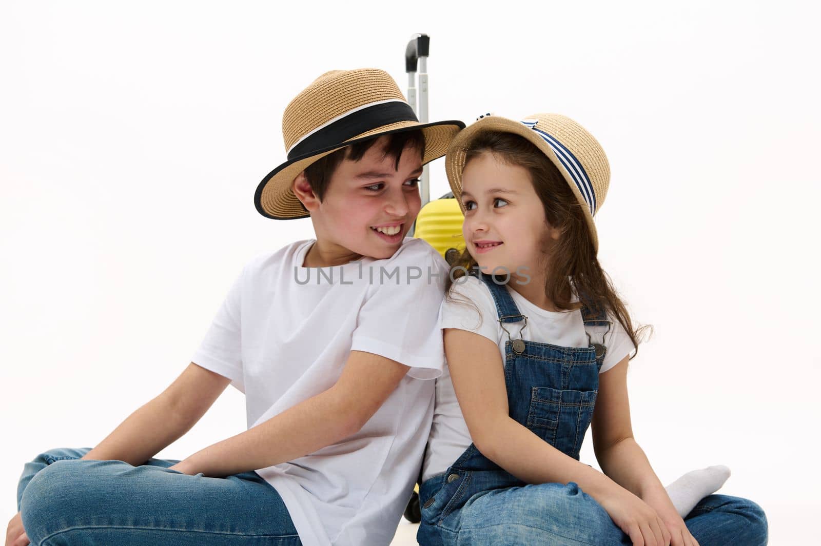 Cute kids, a boy and girl travelers in casual t-shirt and denim wear, with suitcase smile look at each other, on white background. Free ad space. Travel Journey Trip Tourism. Summer vacations concept