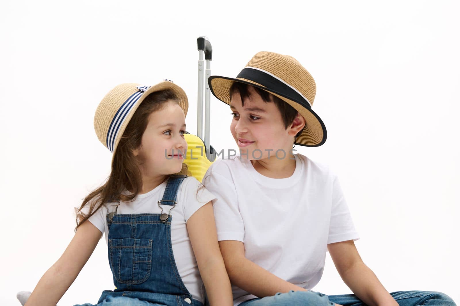 Selective focus on happy travelers kids with yellow suitcase, boy and girl smiling looking at each other, enjoying the upcoming travel abroad for summer holidays, isolated white background. Ad space