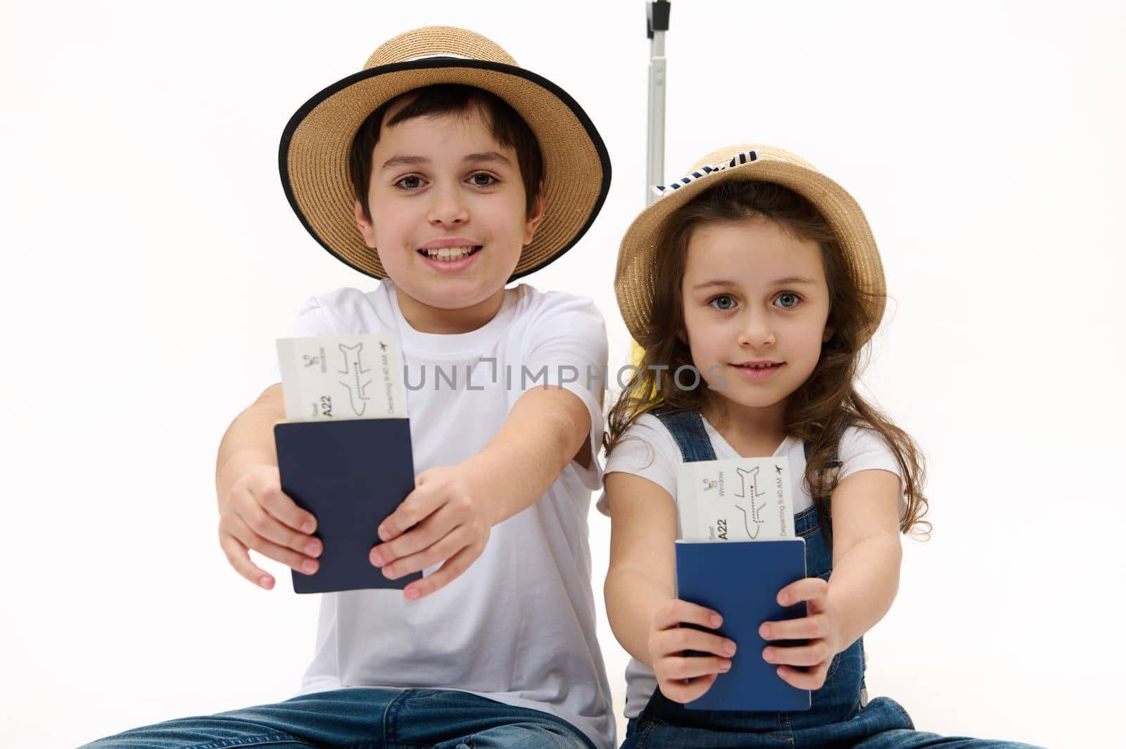 Happy journey. Family of travelers brother and sister wearing casual denim, white t-shirt and straw hat, with suitcase, showing on camera air tickets and passports, isolated on white background